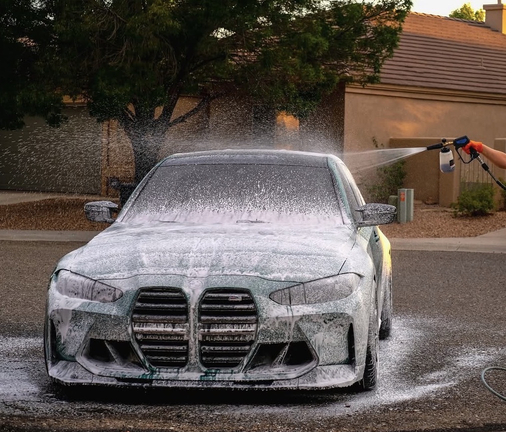 Dark-green BMW M3, foam cannon mid-blast, sunlit